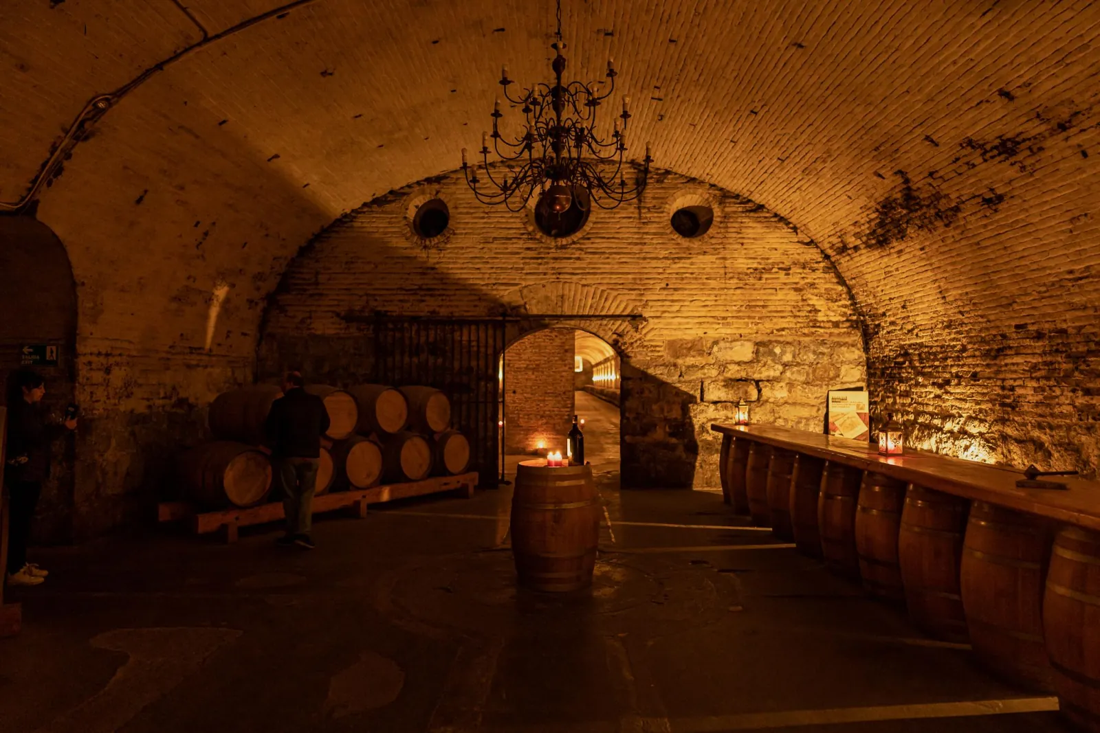Candlelit brick cellar vault with a chandelier, barrels and a long barrel bar