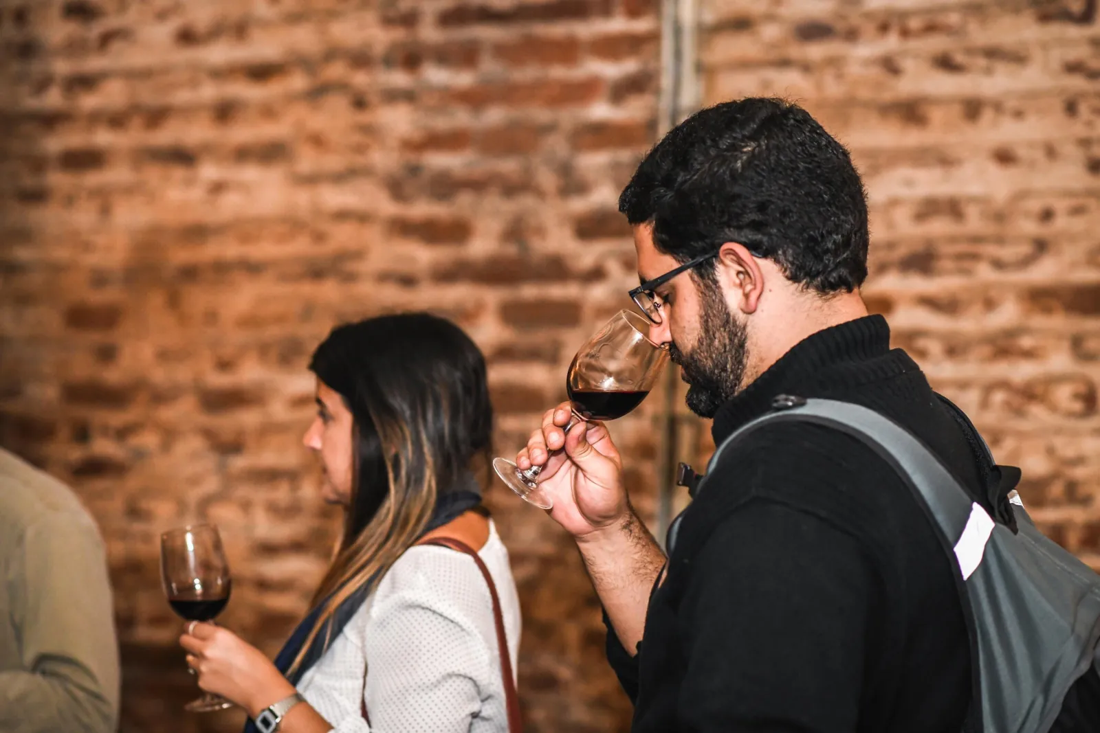 Man smelling red wine beside a woman holding a glass against brick