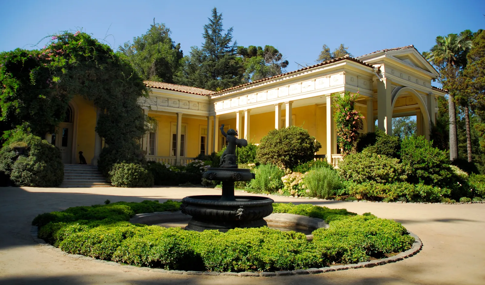Yellow colonnaded mansion behind a dark fountain and round clipped hedge