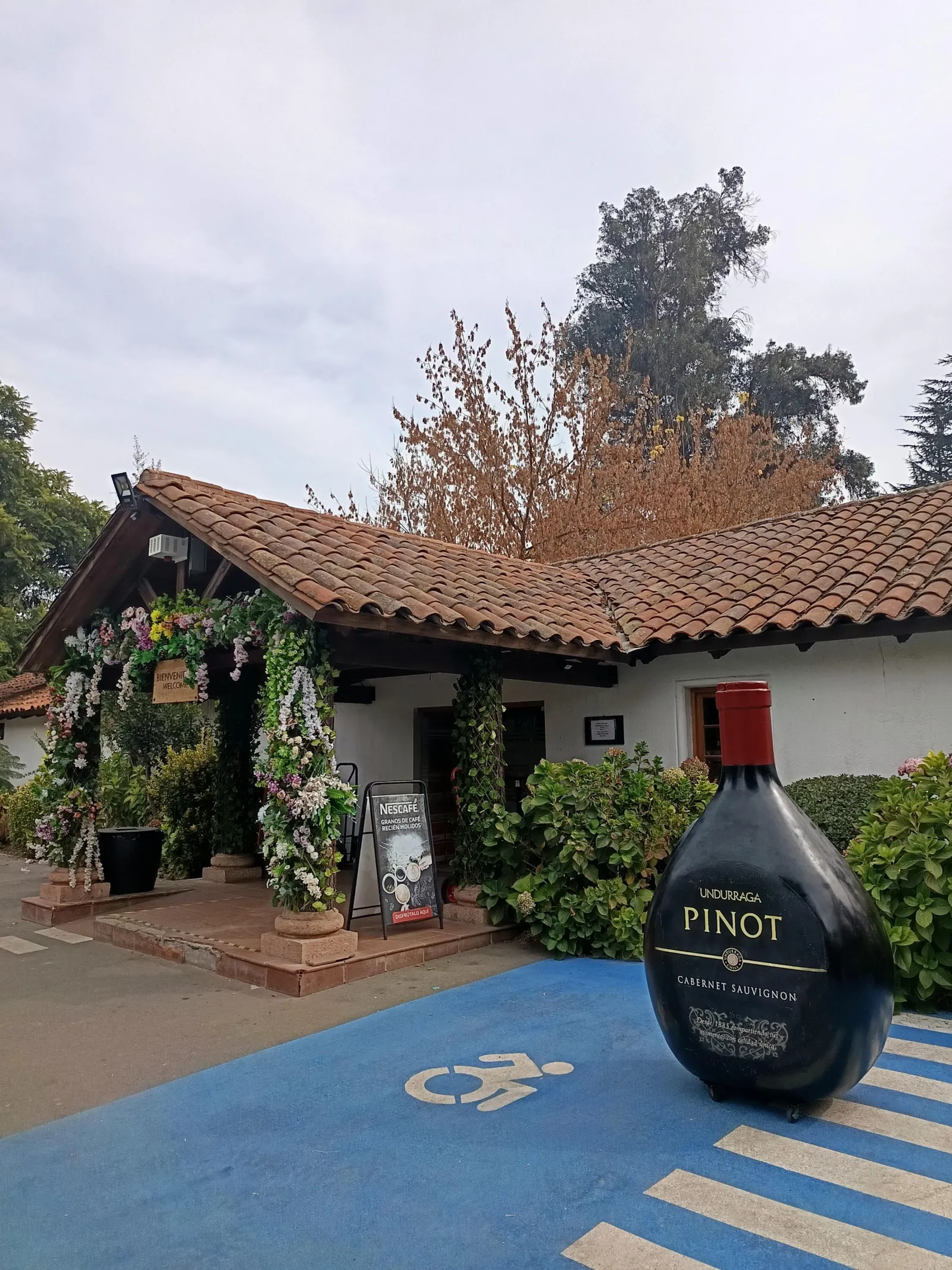Flower-framed entrance of a tiled-roof building beside a giant Undurraga bottle