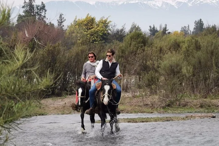 Two riders crossing a stream with snow-capped Andes behind