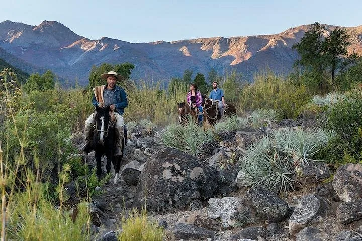 Guide in a wide hat leading two riders along a rocky trail below mountains