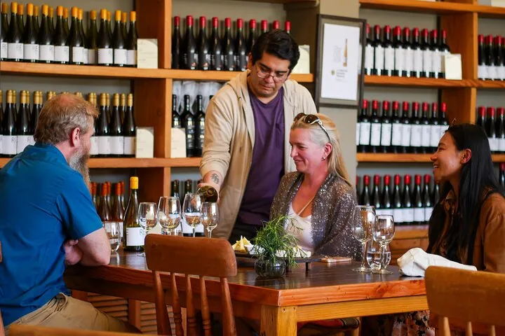 Host pouring wine for guests at a wooden table before bottle shelves