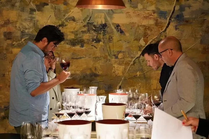Tasters swirling red wine at a table of glasses against a painted wall